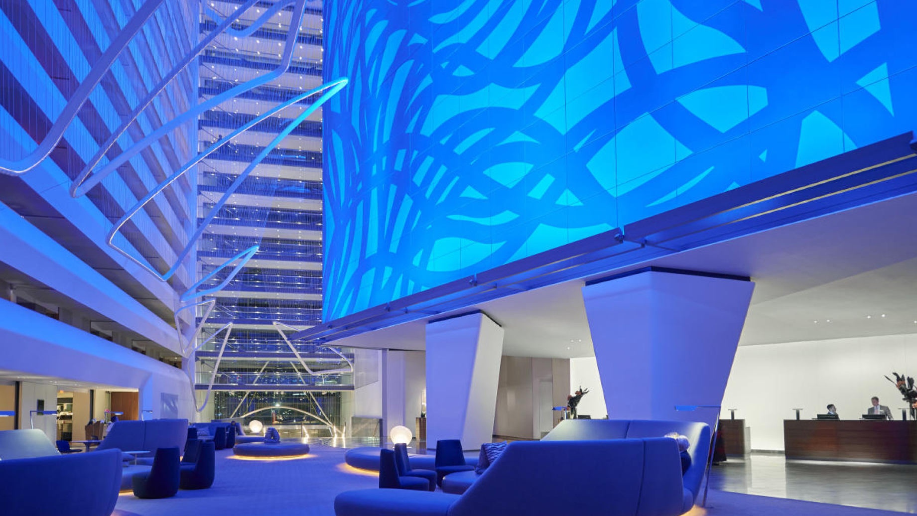 A spacious modern hotel lobby with blue lighting, abstract wall designs, and various seating areas. A reception desk is visible on the far right.