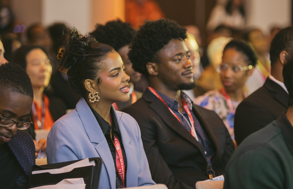 Audience members sitting and listening attentively during an indoor event, wearing badges and business attire.