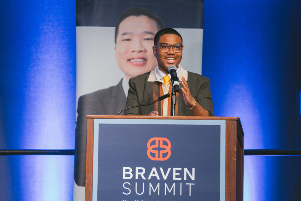 A speaker stands at a podium labeled "Braven Summit," gesturing while speaking, with a large portrait of another person displayed in the background.