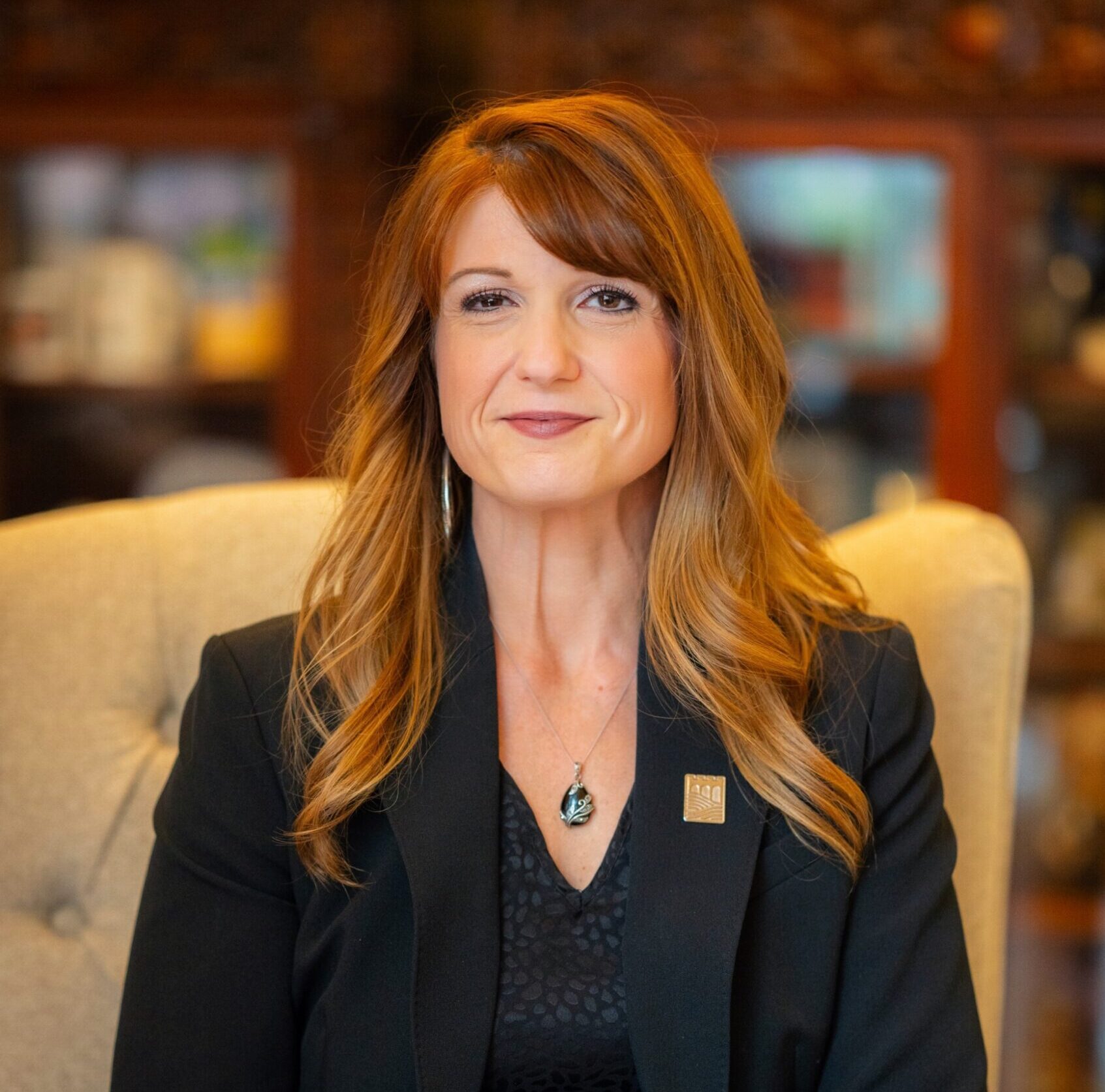 A woman with long, light brown hair sits on a beige chair, wearing a black blazer and top, with her hands folded in her lap, looking at the camera.